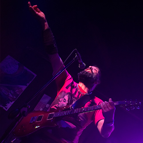 Foto de un cantante frente al micrófono, alzando una mano y con la otra agarrando su guitarra